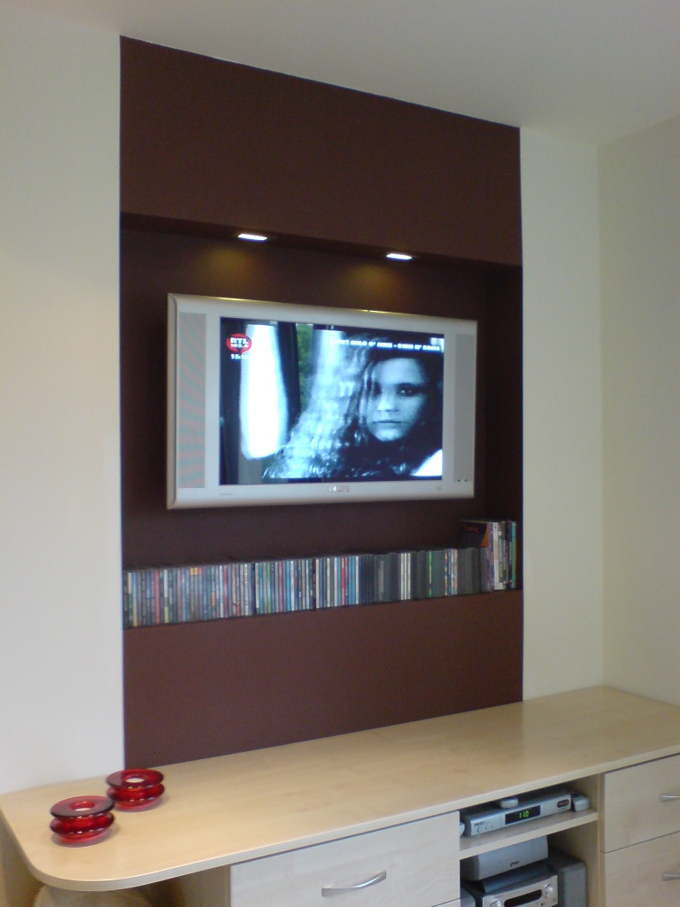 Telewizor LCD zawieszony na brązowej ścianie z podświetleniem LED, poniżej rząd płyt CD i DVD, na szafce RTV czerwone świeczniki.