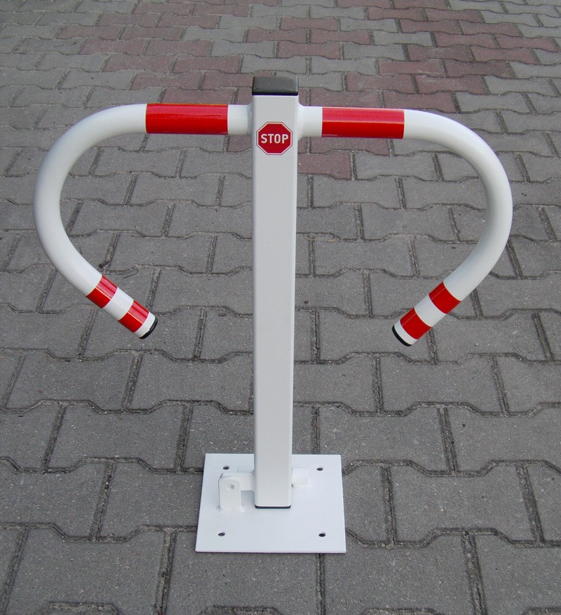 Składana, biała blokada parkingowa z czerwonymi elementami odblaskowymi i znakiem STOP, zamontowana na kostce brukowej.