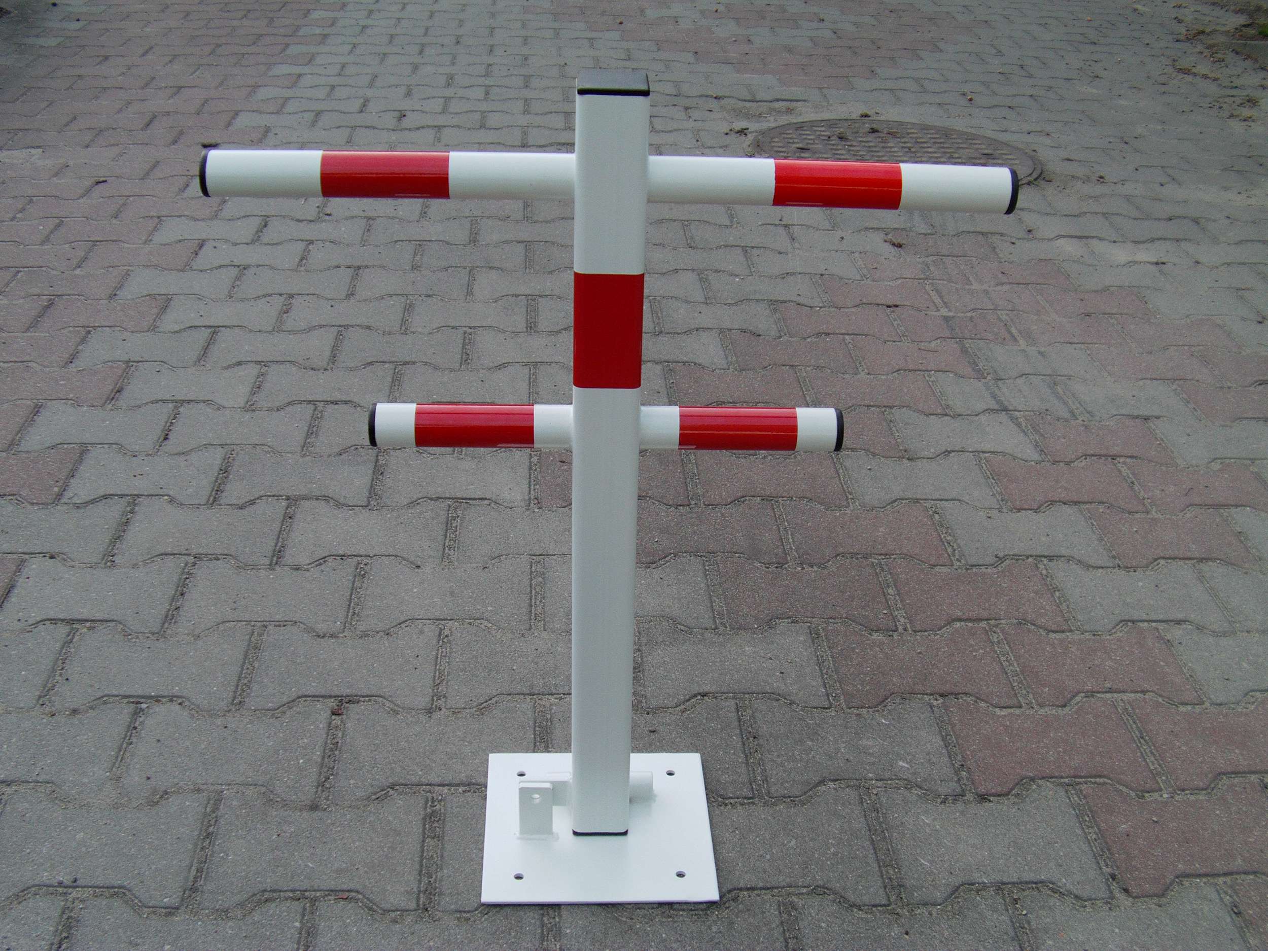 Składana blokada parkingowa w kolorze białym z czerwonymi pasami, zamontowana na kostce brukowej, widoczne elementy mocujące do podłoża.
