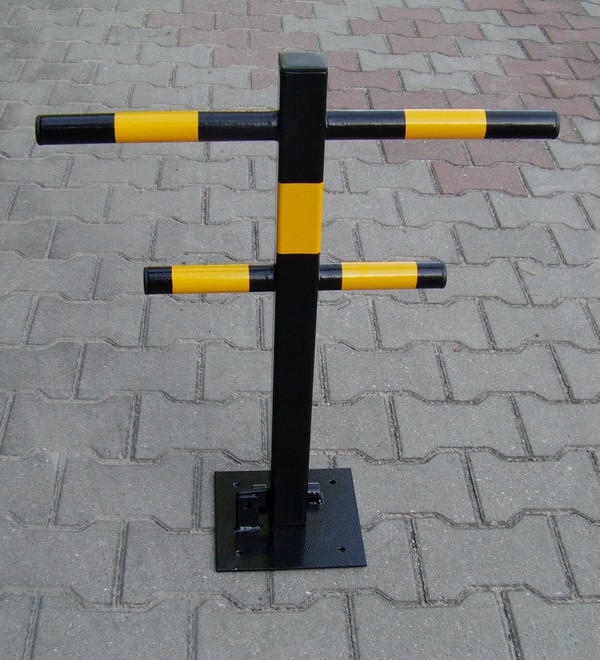 Składana blokada parkingowa w kolorze czarnym z żółtymi pasami ostrzegawczymi, zamontowana na kostce brukowej.