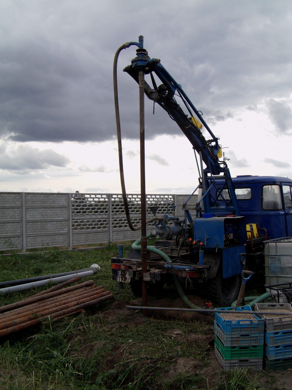 Mobilna wiertnica studni głębinowych na tle pochmurnego nieba i betonowego ogrodzenia, widoczne rury wiertnicze i skrzynki narzędziowe.