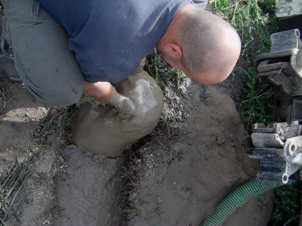 Mężczyzna w trakcie wiercenia studni, ręcznie wybiera błoto z otworu, obok widoczny fragment wiertnicy.