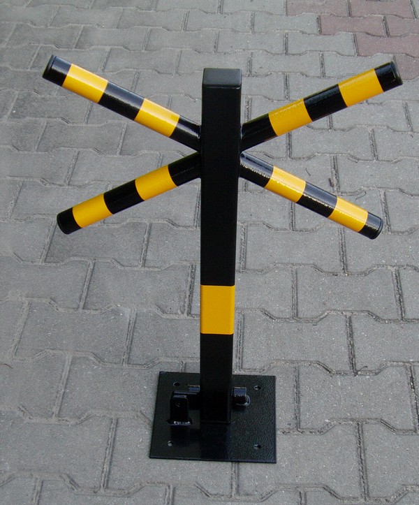 Składana blokada parkingowa w kolorze czarnym z żółto-czarnymi pasami na ramionach, zamontowana na szarej kostce brukowej.