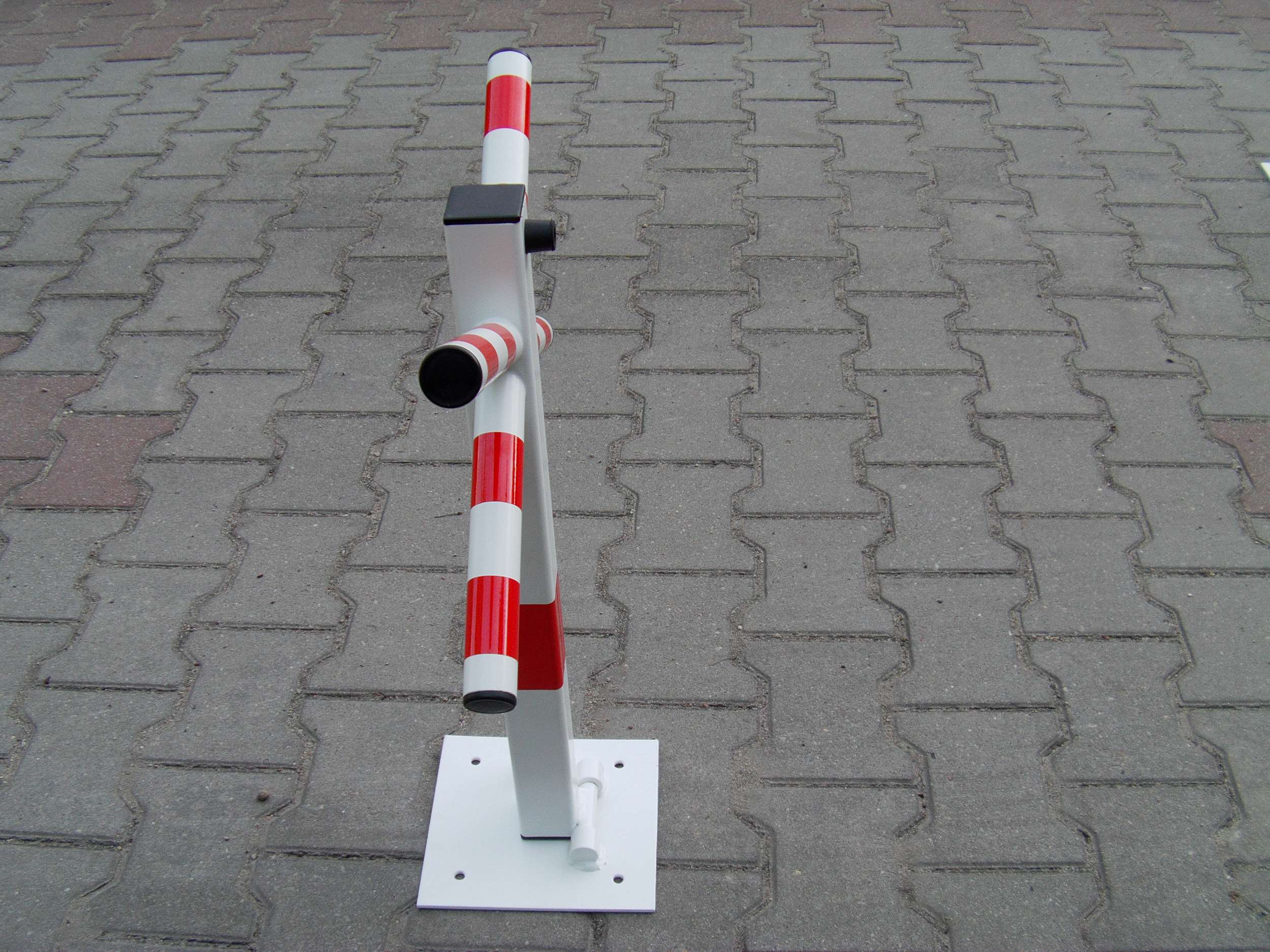 Składana blokada parkingowa w kolorze białym i czerwonym, zamontowana na kostce brukowej, widok z góry.