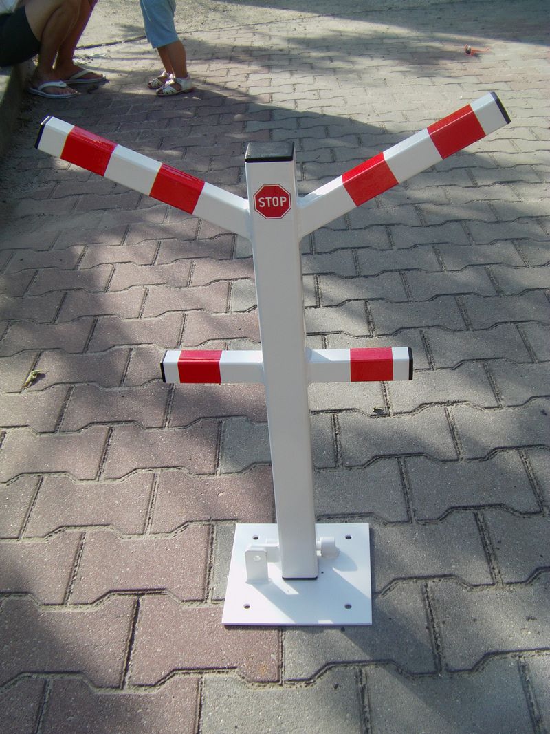 Składana, biała blokada parkingowa z czerwonymi pasami ostrzegawczymi i znakiem STOP, zamontowana na kostce brukowej.