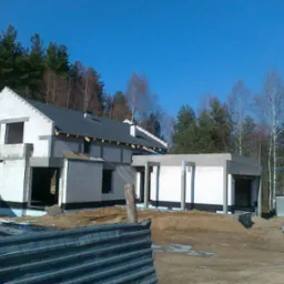 Surowy stan domu z białego bloczka, z częściowo położonym ciemnym dachem, na tle lasu i błękitnego nieba, widoczny fragment ogrodzenia i tablica ostrzegawcza.