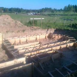 Zalane wykopy pod fundamenty z drewnianym szalunkiem i zbrojeniem, widok z góry na tle zielonego krajobrazu.