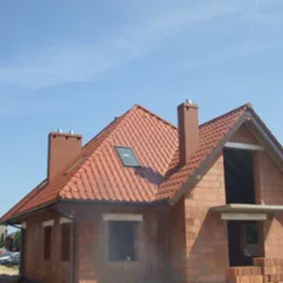 Wznoszony dom jednorodzinny z czerwonej cegły z dachem pokrytym czerwoną dachówką, widoczne kominy i okno dachowe, stan surowy otwarty, błękitne niebo w tle.