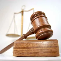 Drewniany młotek sędziowski na podstawie, w tle rozmyte złote wagi sprawiedliwości na białym tle.