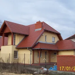 Żółty dom jednorodzinny z czerwoną dachówką, drewnianą konstrukcją werandy i wykończeniami, widoczny zza ogrodzenia z siatki, z datą 17/01/2005 w prawym dolnym rogu.