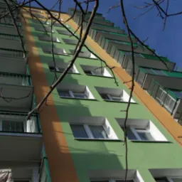 Wielopiętrowy budynek mieszkalny z zieloną elewacją i pomarańczowym pasem, widok z dołu, z gałęziami drzew na pierwszym planie.