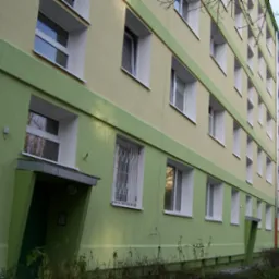 Wielopiętrowy budynek mieszkalny z oknami w białych ramach i zielonymi elementami elewacji, widoczne krzewy u podstawy budynku.