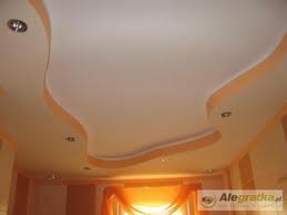 Biały sufit podwieszany z nieregularnymi, łagodnymi krzywiznami, obramowany delikatnym, pomarańczowym paskiem, oświetlony punktowymi lampami halogenowymi.