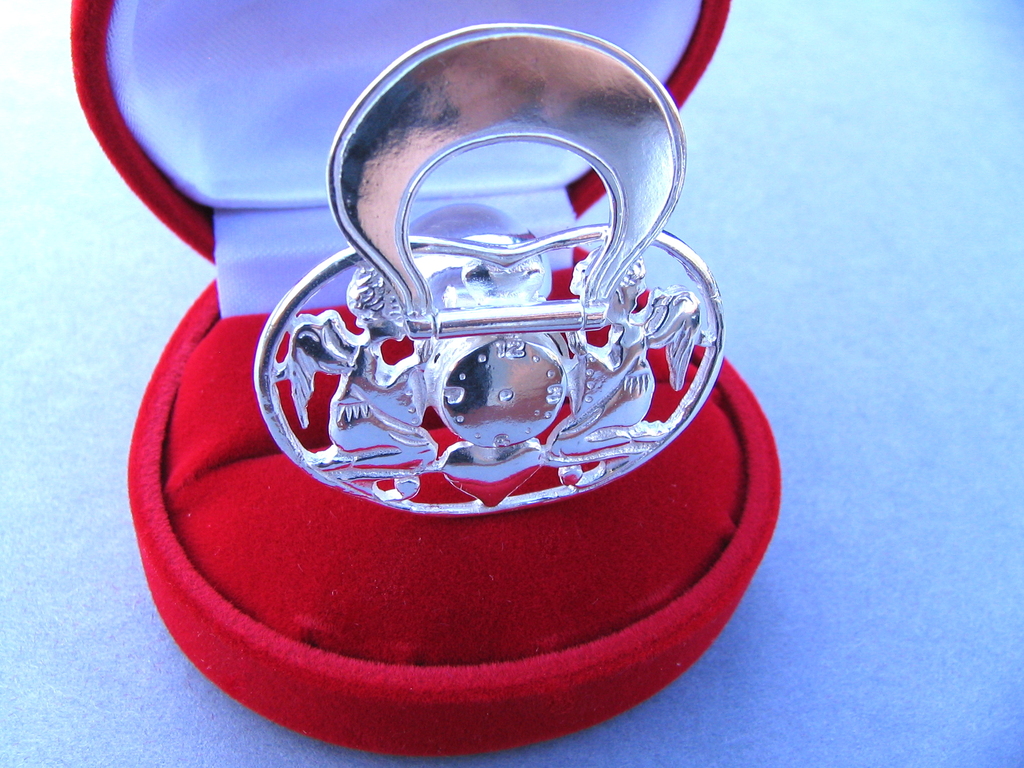 Srebrny smoczek z motywem aniołków i zegara w czerwonym, welurowym pudełku prezentowym.