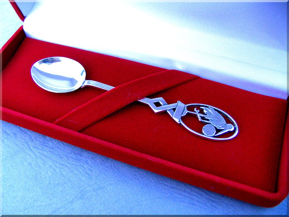 Srebrna łyżeczka z grawerowanym symbolem bociana w owalnej ramce, prezentowana w czerwonym welurowym pudełku z białym wnętrzem.