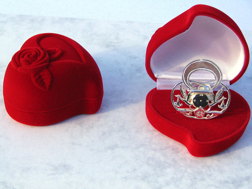 Srebrny smoczek-zegarek z motywem wagi w otwartym, czerwonym, welurowym pudełku w kształcie serca, obok zamknięte pudełko z wytłoczonym motywem róży.