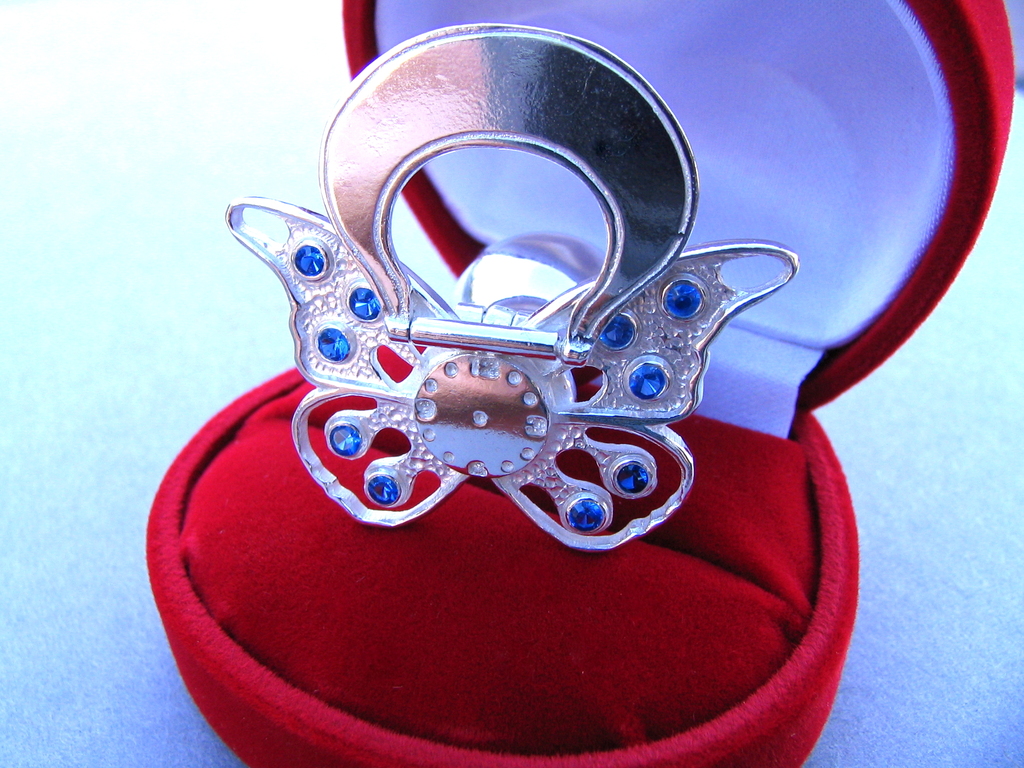 Srebrny smoczek w kształcie motyla z niebieskimi kamieniami w czerwonym, welurowym pudełku.