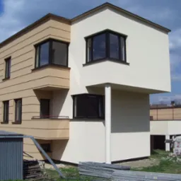 Dwukondygnacyjny dom z elewacją w odcieniach beżu i kremu, z widocznymi elementami rusztowania i materiałów budowlanych na trawniku.