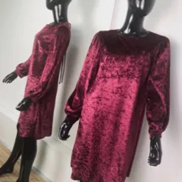 Dwie błyszczące, czarne manekiny prezentują bordowe, welurowe sukienki z długimi, bufiastymi rękawami na tle białej ściany, z widocznym fragmentem dywanu w dolnym rogu.