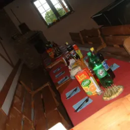 Przygotowania do imprezy w surowym wnętrzu: stół nakryty czerwonym obrusem, napoje, przekąski, w tle widoczny kominek z cegły i drewniane ławy.