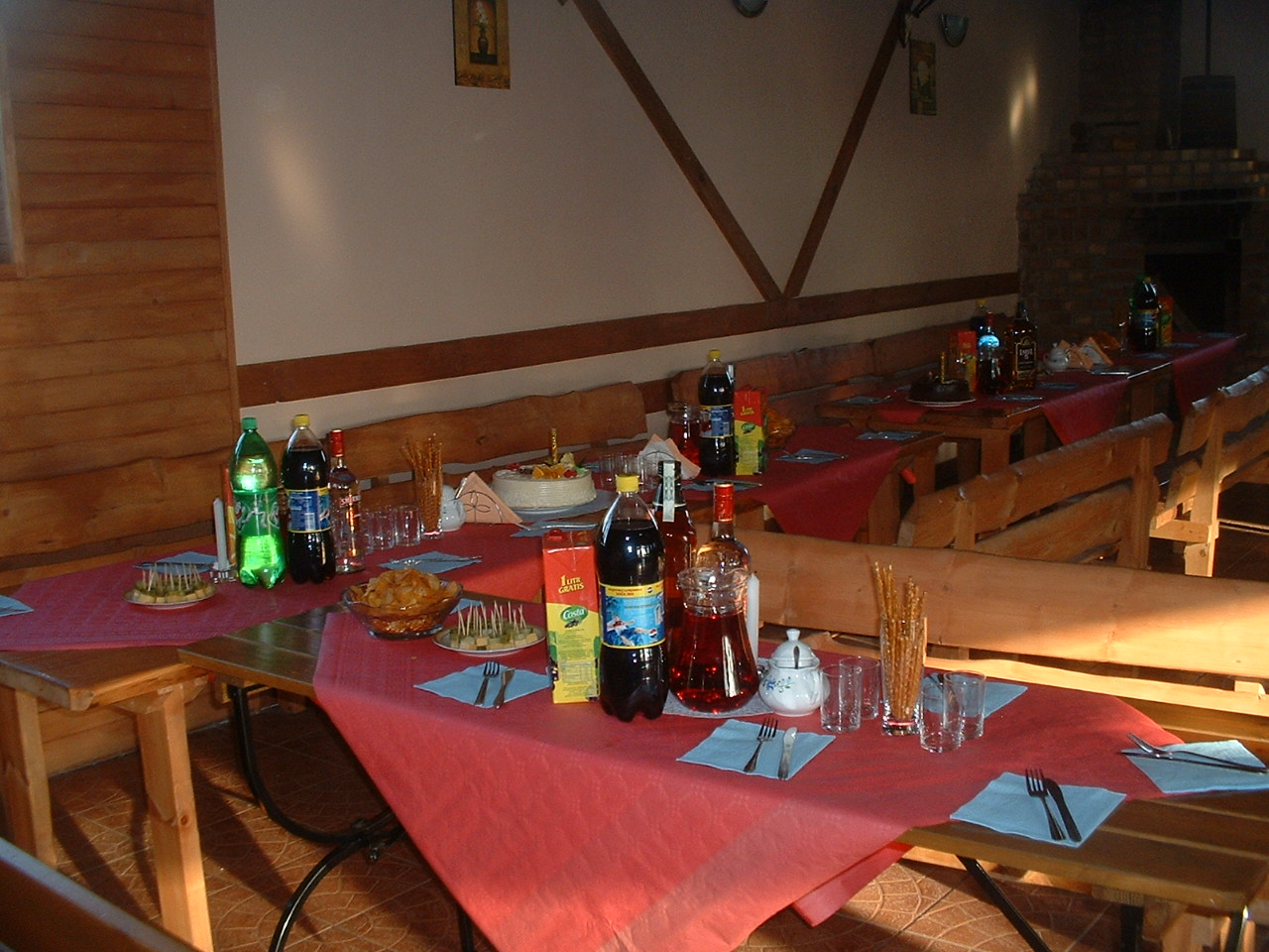 Ustawione stoły z napojami, przekąskami i ciastem w drewnianej sali z ławami, przygotowane na imprezę.