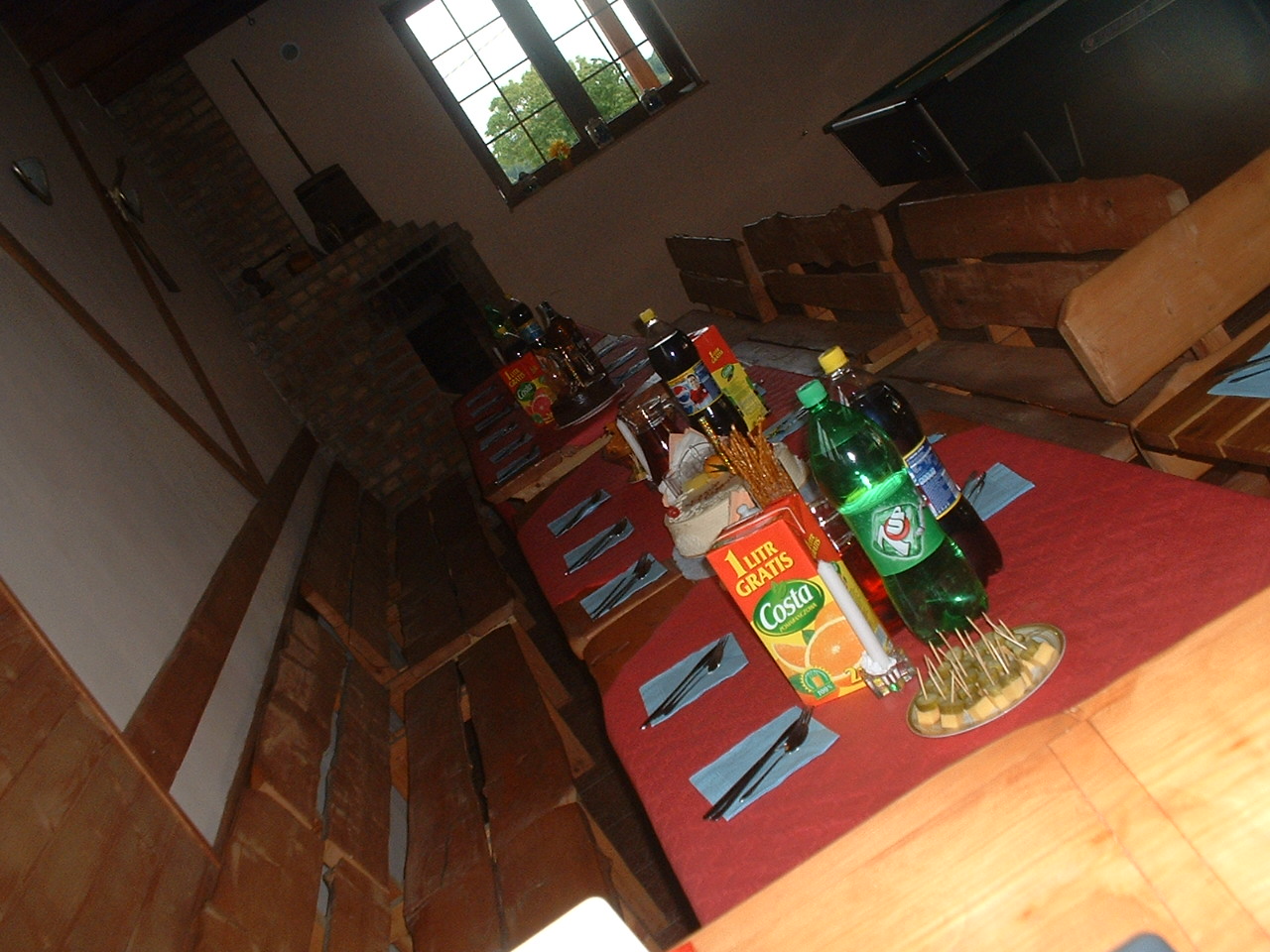 Przygotowania do imprezy w surowym wnętrzu: stół nakryty czerwonym obrusem, napoje, przekąski, w tle widoczny kominek z cegły i drewniane ławy.