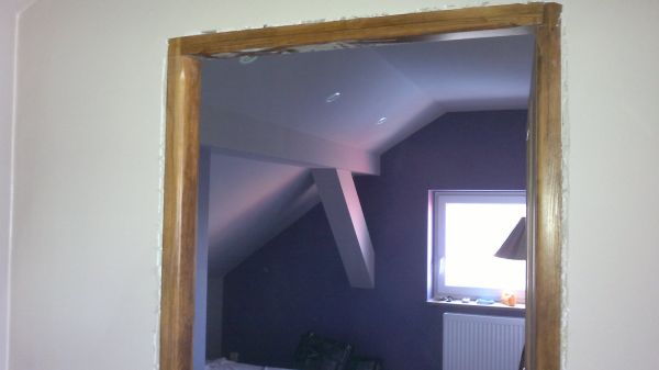 Drewniana ościeżnica drzwiowa w trakcie montażu, widoczna szpara między ościeżnicą a ścianą, w tle pokój na poddaszu z oknem i grzejnikiem.