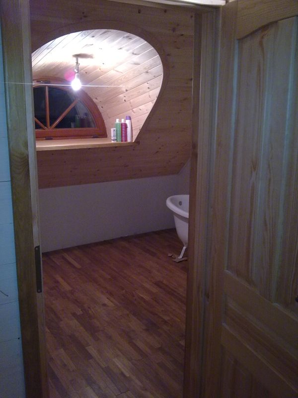 Drewniane wykończenie łazienki na poddaszu z nietypowym oknem w kształcie łzy, widok zza uchylonych drzwi.