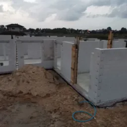 Wznoszone ściany parteru domu z białych bloków silikatowych, widoczny fundament, drewniane szalunki i sterta piasku na placu budowy, w tle zabudowa jednorodzinna pod pochmurnym niebem.