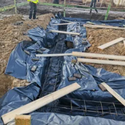 Przygotowanie wykopu pod fundament zbrojonego, zabezpieczonego folią, z widocznymi deskami szalunkowymi i pracownikami w oddali.