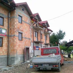 Budynek w trakcie budowy, widoczne okna, instalacja elektryczna zewnętrzna, reklama firmy Westplast oraz samochód dostawczy z piaskiem.