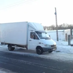 Biały samochód dostawczy Mercedes Sprinter z zabudową kontenerową, zaparkowany na ośnieżonej drodze w zimowy dzień, widoczne budynki gospodarcze w tle.