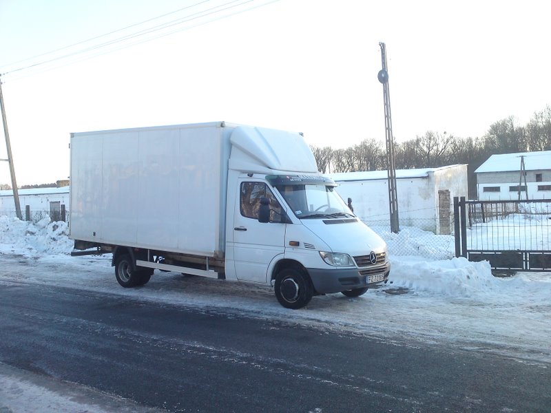 Biały samochód dostawczy Mercedes Sprinter z zabudową kontenerową, zaparkowany na ośnieżonej drodze w zimowy dzień, widoczne budynki gospodarcze w tle.