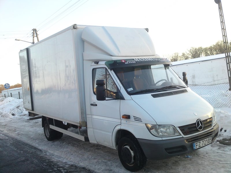 Biały samochód dostawczy Mercedes-Benz Sprinter z zabudową kontenerową, zaparkowany na zaśnieżonej ulicy, z widoczną naklejką 'MAXIMUS' nad przednią szybą.