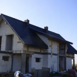 Dom w stanie surowym otwartym z ciemną dachówką i widocznymi kominami. Elementy konstrukcyjne z drewna. Budowa w toku, w słoneczny dzień.