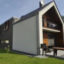 Dwukondygnacyjny dom z balkonem, elewacją z tynku i cegły, widoczny grill i meble ogrodowe na trawniku. Dach z anteną satelitarną.