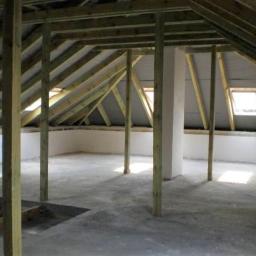 Stamann - Surowe poddasze z drewnianą konstrukcją dachu, widoczne okna dachowe i betonowa podłoga w trakcie prac wykończeniowych.