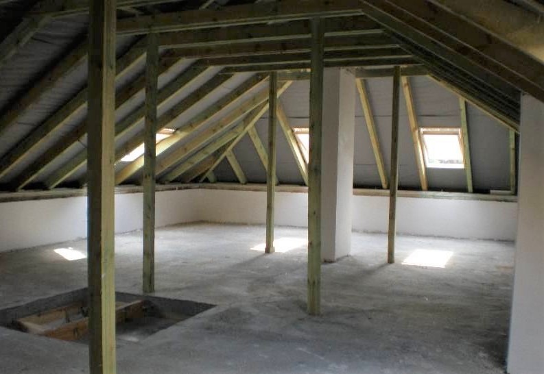 Surowe poddasze z drewnianą konstrukcją dachu, widoczne okna dachowe i betonowa podłoga w trakcie prac wykończeniowych.