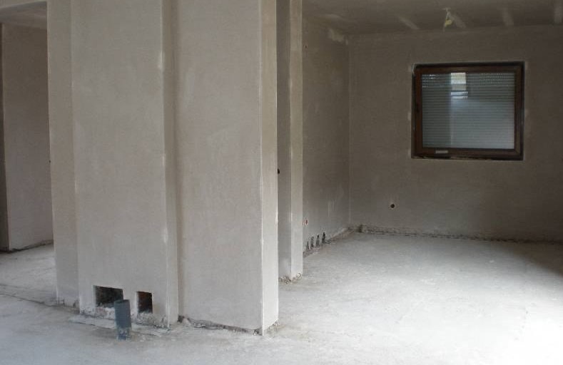 Surowe wnętrze w trakcie prac wykończeniowych: ściany pokryte szarą, nierówną warstwą, widoczne otwory instalacyjne w kolumnie, okno z żaluzjami w tle, surowy beton na podłodze.