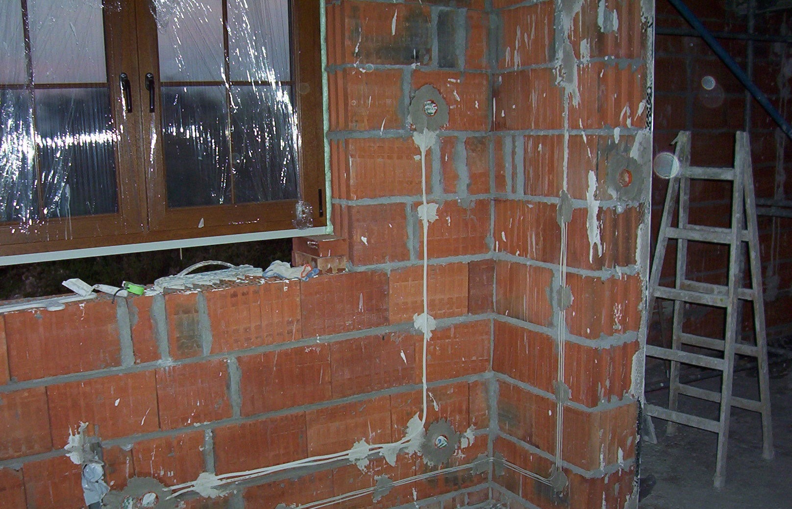 Surowa ściana z czerwonej cegły z widocznymi przewodami elektrycznymi i puszkami instalacyjnymi, okno zabezpieczone folią, drabina w tle.