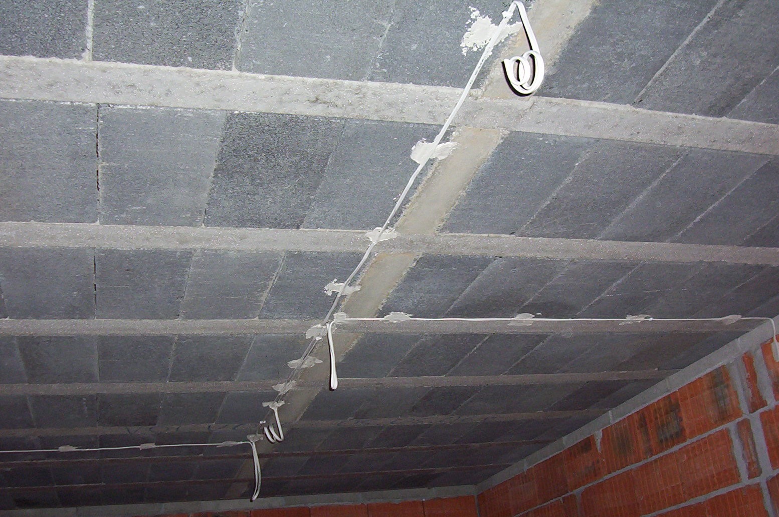 Betonowy strop z widocznymi przewodami elektrycznymi przymocowanymi do sufitu za pomocą uchwytów i gipsu, ściany z czerwonej cegły w tle.