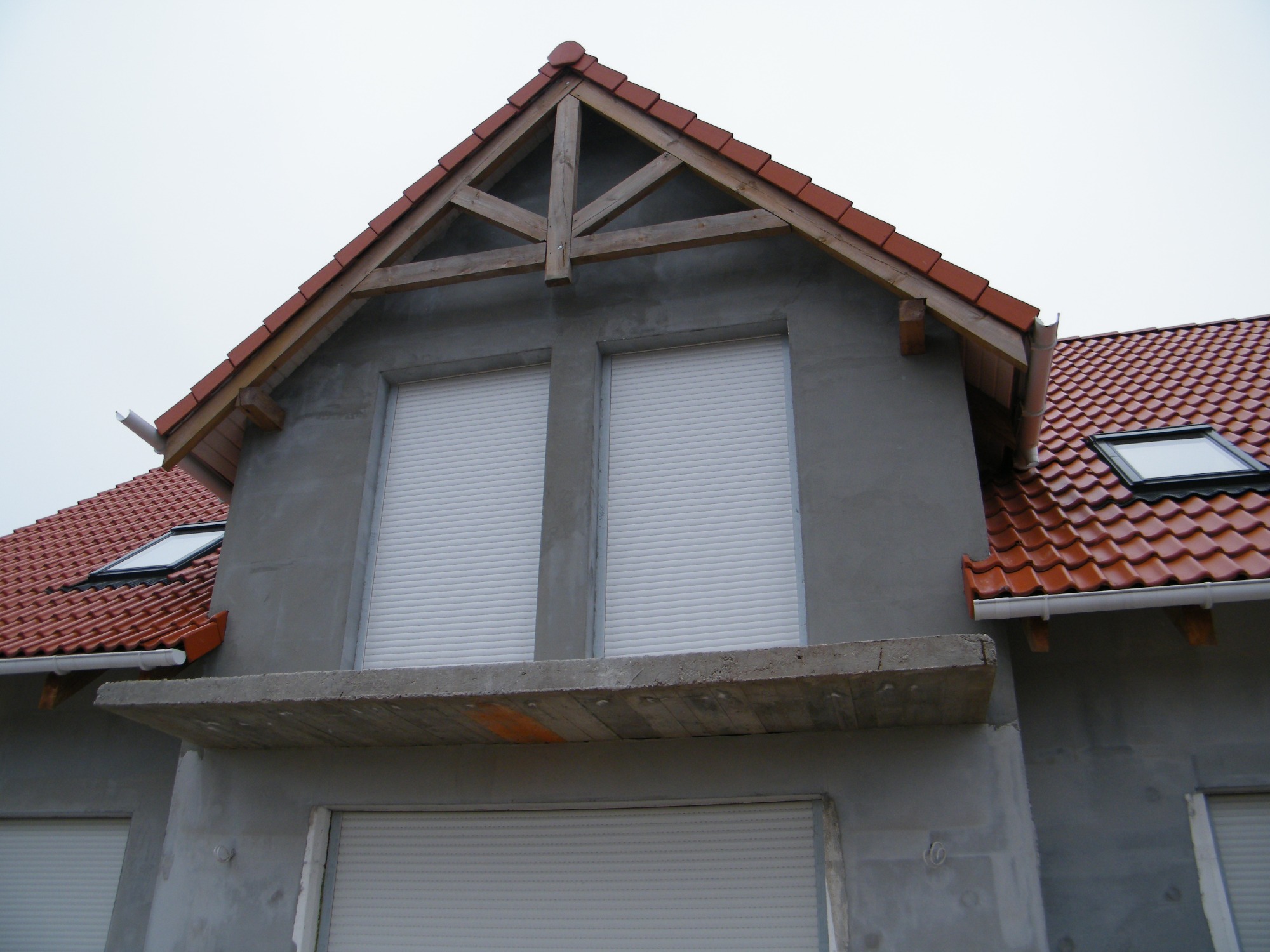 Surowy stan elewacji domu z widoczną konstrukcją dachu, oknami z roletami zewnętrznymi i betonowym balkonem w trakcie budowy.