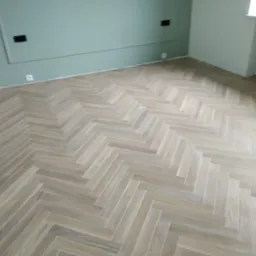 Parkiet ułożony w jodełkę na podłodze w pokoju z zielonymi i białymi ścianami, widoczne gniazdka elektryczne i grzejnik pod oknem.