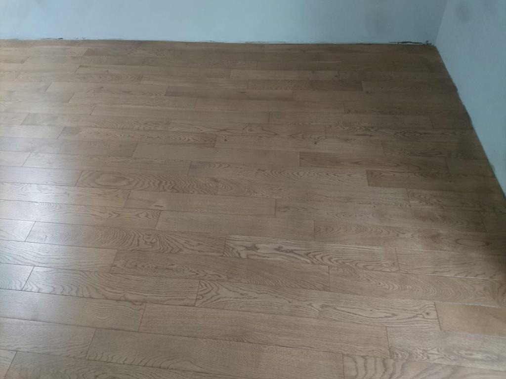 Ułożona, drewniana podłoga w odcieniach brązu, przy białej ścianie, widoczne niedociągnięcia przy listwach przypodłogowych.