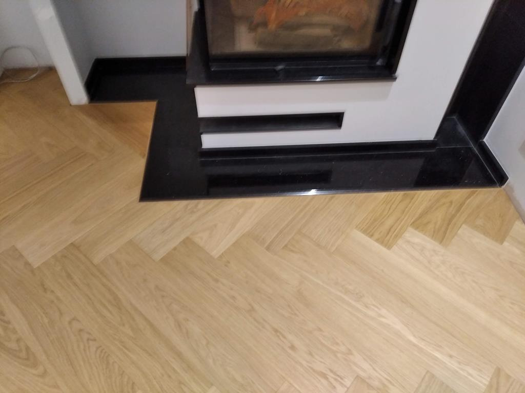 Jasny parkiet ułożony we wzór jodełki francuskiej, przylegający do czarnego, błyszczącego obramowania kominka z białymi elementami dekoracyjnymi.