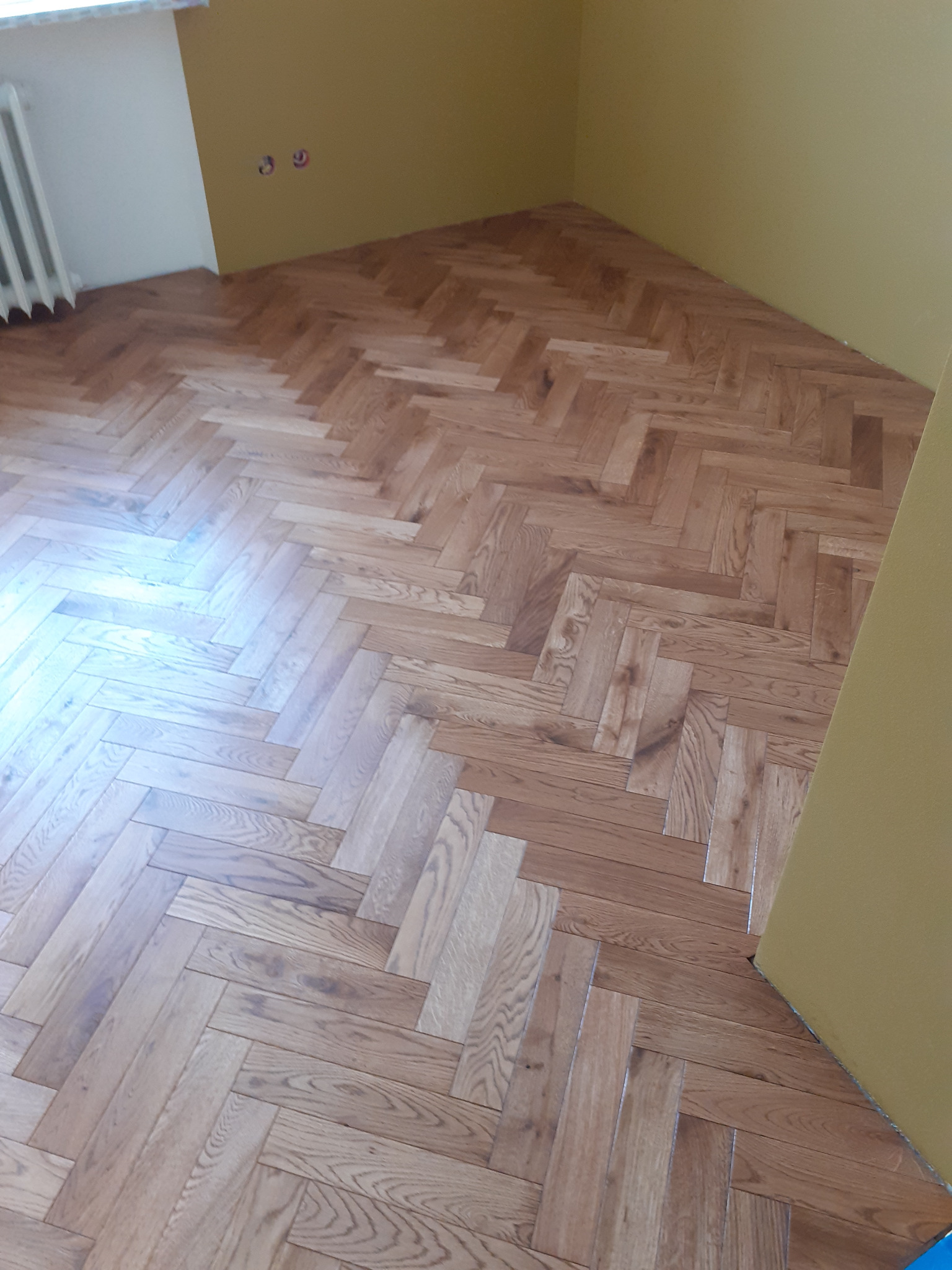 Parkiet ułożony w jodełkę w rogu pokoju, przy żółtej ścianie, widoczny grzejnik.