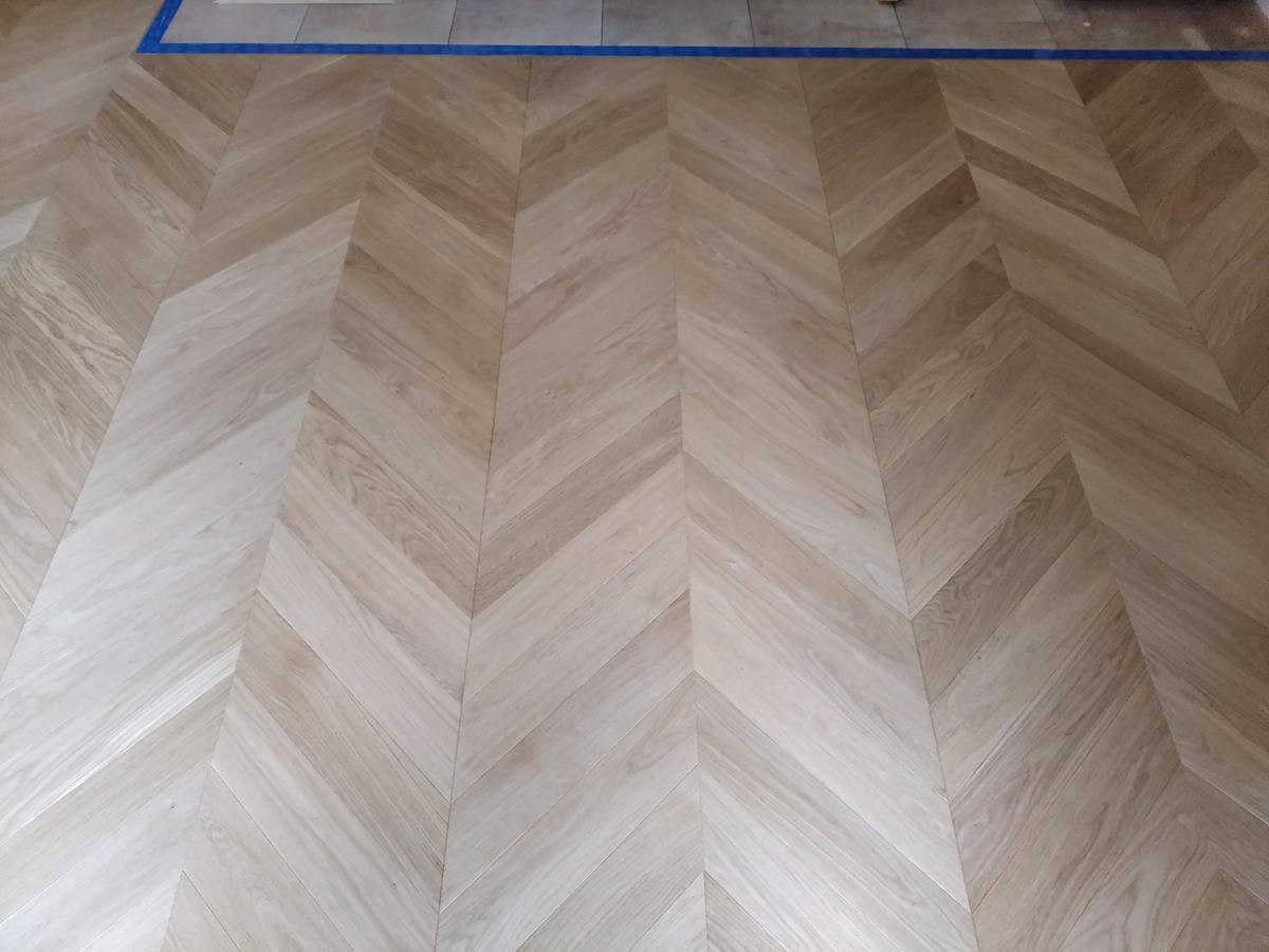 Jasna, dębowa podłoga ułożona we wzór jodełki francuskiej, zabezpieczona niebieską taśmą malarską przy ścianie.