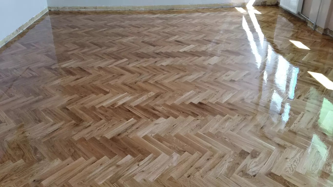 Odnowiona podłoga parkietowa ułożona we wzór jodełki, świeżo polakierowana, z widocznym połyskiem i odbiciem światła słonecznego wpadającego przez okno.