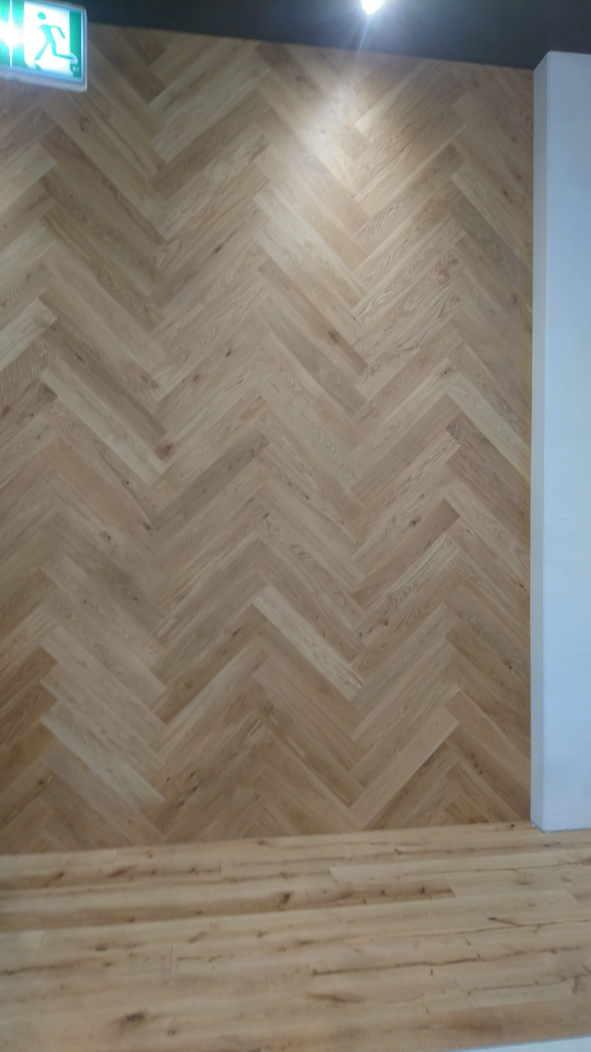 Ściana i podłoga wyłożone parkietem w jodełkę, oświetlone punktowym światłem, z widocznym znakiem ewakuacyjnym w lewym górnym rogu.