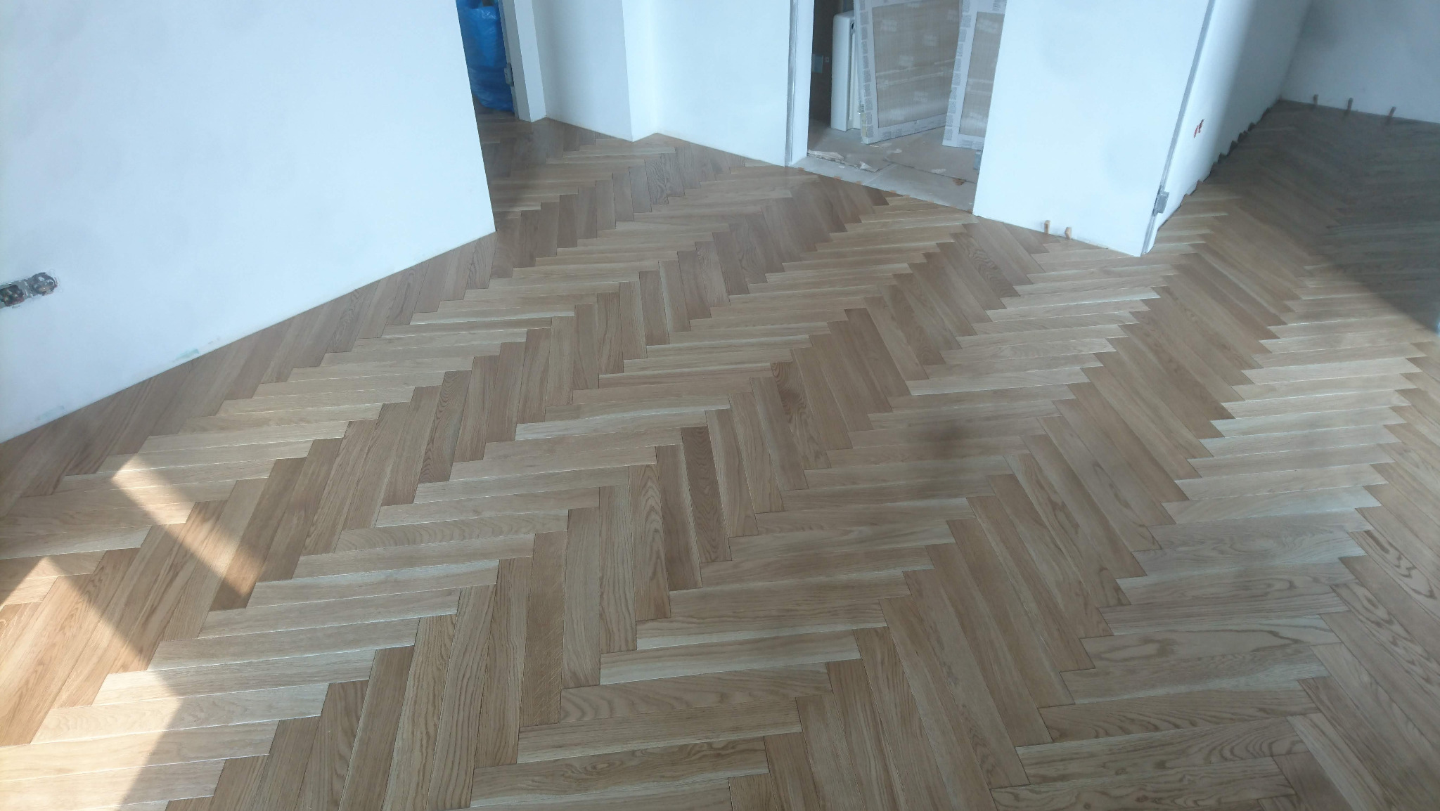 Ułożona parkietowa podłoga w jodełkę, jasne drewno, widoczne fragmenty ścian i drzwi w stanie surowym.